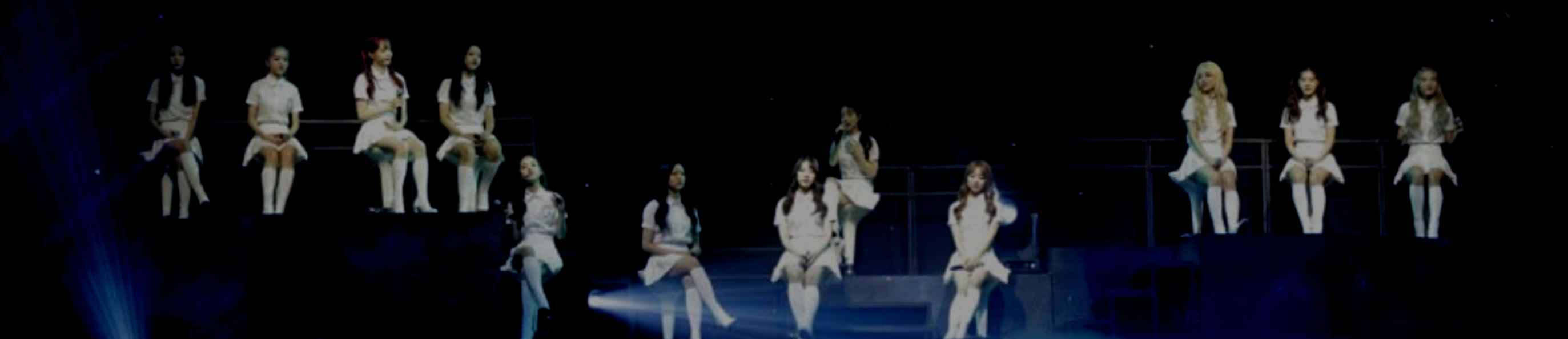 A cropped picture from the Loonabirth concert. The members of Loona are sitting in chairs on a dark stage, illuminated by soft light. They are all wearing white shirts, skirts, socks, and shoes. The members of yyxy are sitting on the left, 1/3 are sitting lower in the middle, with Yeojin sitting right above them in between them, and Odd Eye Circle are sitting up on the right.