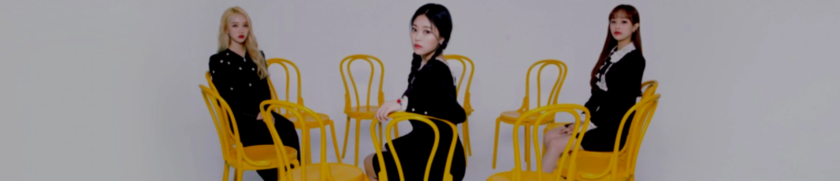 A cropped version of the promotional image for the Premier Greeting: Meet and Up event. From left to right: Go Won, Hyunjin, Chuu. They are sitting in a circle of twelve yellow chairs, with all the other chairs empty. They are looking out at the viewer. They are all wearing black dresses. Go Won has long blonde hair, Hyunjin has long black hair styled into two braided pigtails, and Chuu has long reddish brown hair. The background is a white void.