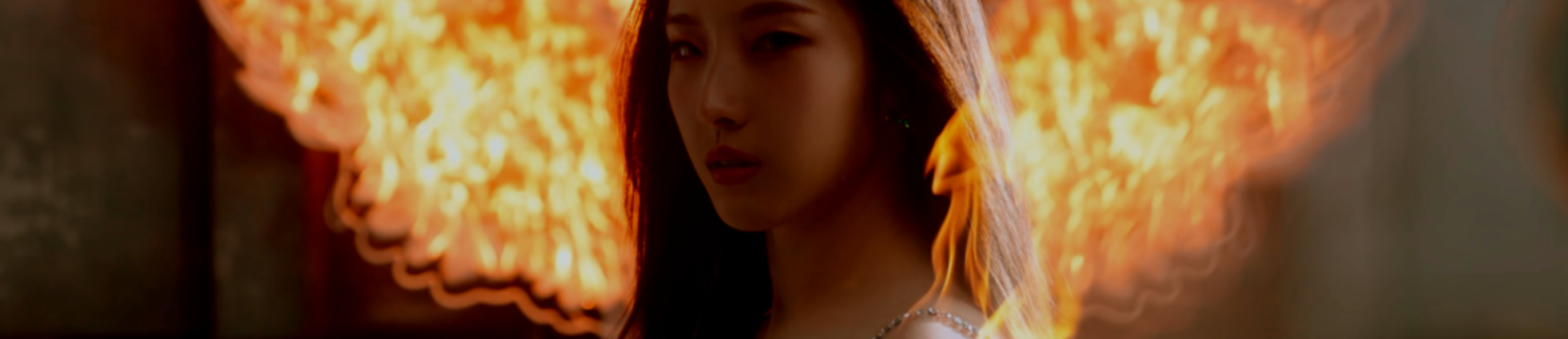 A cropped screenshot from the Paint the Town music video. Haseul is standing in the center. She is facing the left, with her face turned to look at the viewer. She has long brown hair. Her shoulder is on fire, but she has a neutral expression. There are LED lights in the shape of wings behind her, also on fire.