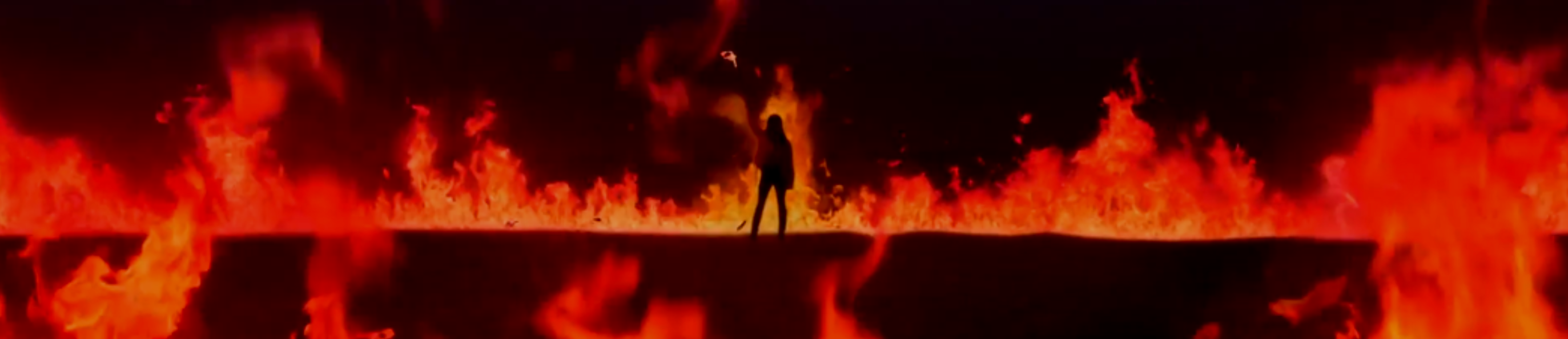 A cropped screenshot from the So What music video. The silhouette of Olivia Hye is standing far back in the middle, holding up a torch on fire. She is in a dark landscape, and there is bright fire burning all around her.