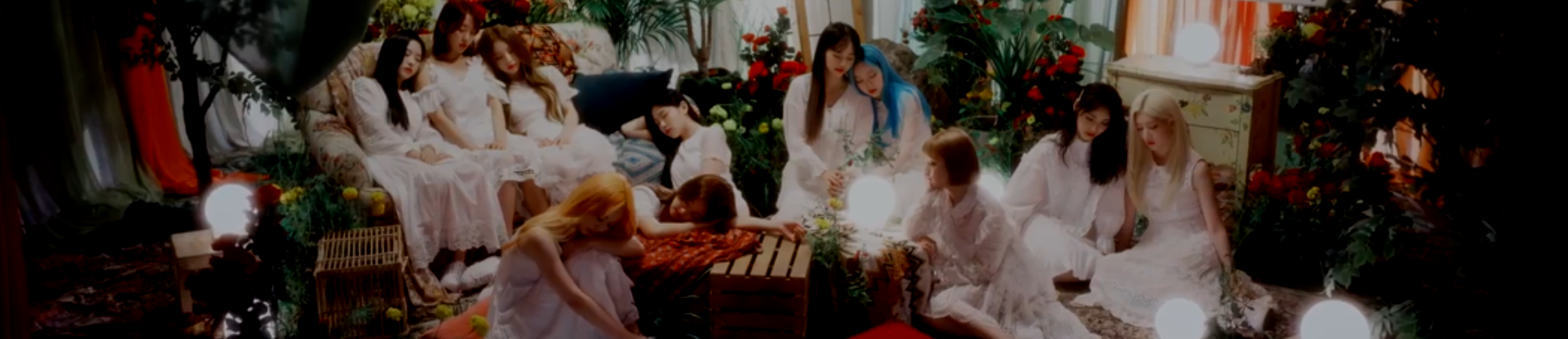 A cropped screenshot from the Star music video. The members of Loona, minus Haseul, are asleep in a very decorated room. They are all wearing lacy white dresses. The room is full fo furniture, including couches and tables, with lots of plants and flowers surrounding them. There are also glowing moon-like orbs all around the room, and curtains in the back.