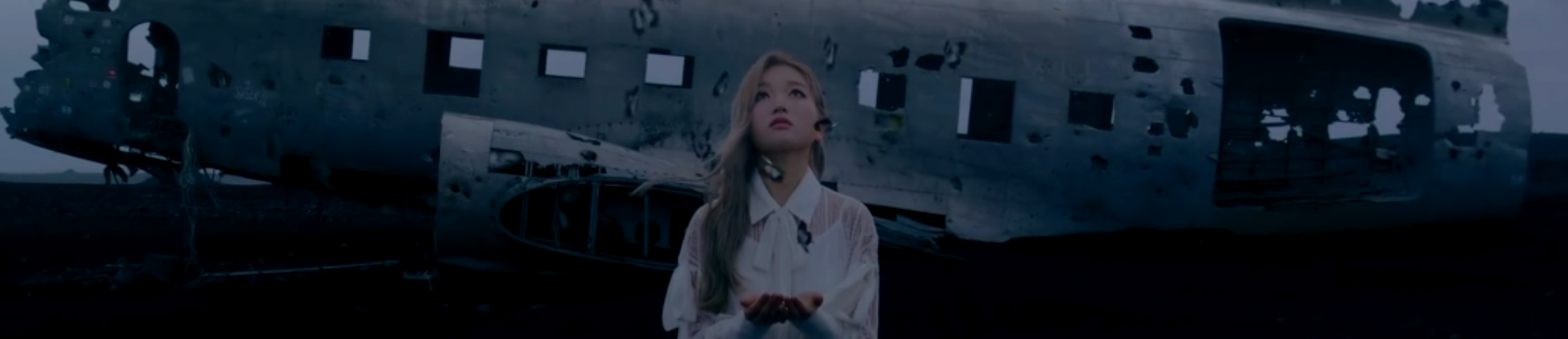 A cropped screenshot from the XIIX teaser. Go Won is standing in front of the crashed plane from the Let Me In music video, in the gray and black Iceland landscape. She is holding her hands out in front of her and looking up at the multiple butterflies flying up from her hands. She has long blonde hair, and is wearing a lacy white dress.