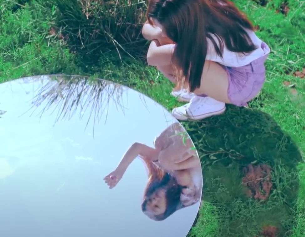 A screenshot from the Love Cherry Motion music video. Choerry is wearing a white shirt with light purple shorts and white Converse. She has long dark brown hair. She is in a green grassy field, crouching in front of a large circular mirror on the ground. In the reflection, another Choerry, wearing the same outfit and crouching in the same way, is holding a cherry by the stem out in front of her.