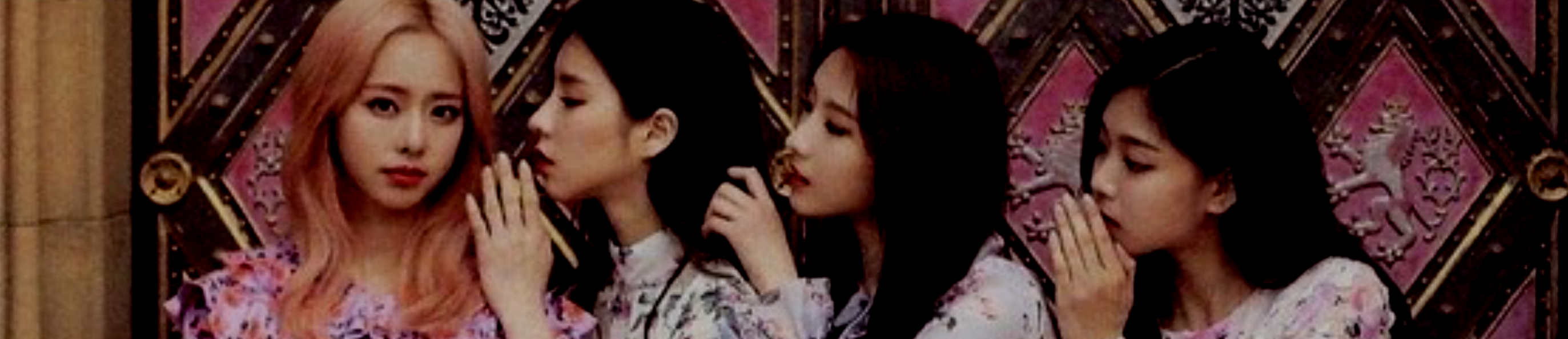 A cropped image of the members of Loona 1/3. From left to right: Vivi, Heejin, Haseul, Hyunjin. They are wearing frilly dresses with floral patterns. Vivi has long light pink hair and is looking at the viewer with a half smile. The other members have cupped their hands and are whispering into each other's ears in a row, like a game of telephone. Heejin has long black hair, Haseul has long brown hair, and Hyunjin has long black hair. They are sat in front of a large gold and magenta door.