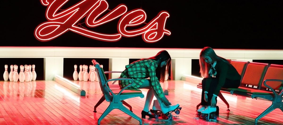 A cropped image of Yves and Vivi from the new music video. They are sat on benches facing each other, in front of a small bowling alley. They are illuminated by bright green and red lights. Yves is wearing her yellow plaid yyxy uniform, while Vivi wears a green sweater and black pants. Yves has long black hair, and Vivi has long blonde hair. They are both in the process of putting on white roller skates. Behind them are bowling lanes, with neon lights and pins set up in each lane. There is a large LED sign on the wall that reads 'Yves' in red cursive text. The wall is black.