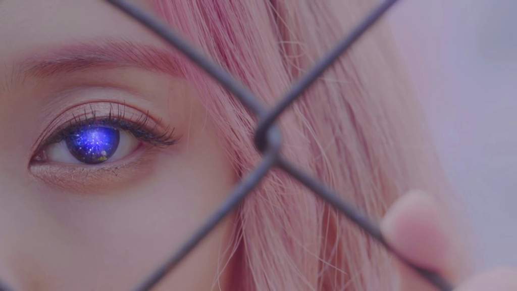 A close up of ViVi's left eye from the Love & Live music video. Her pupil is purple and black with twinkling stars, resembling a galaxy. She has light pink hair. There is a fence in front of her.