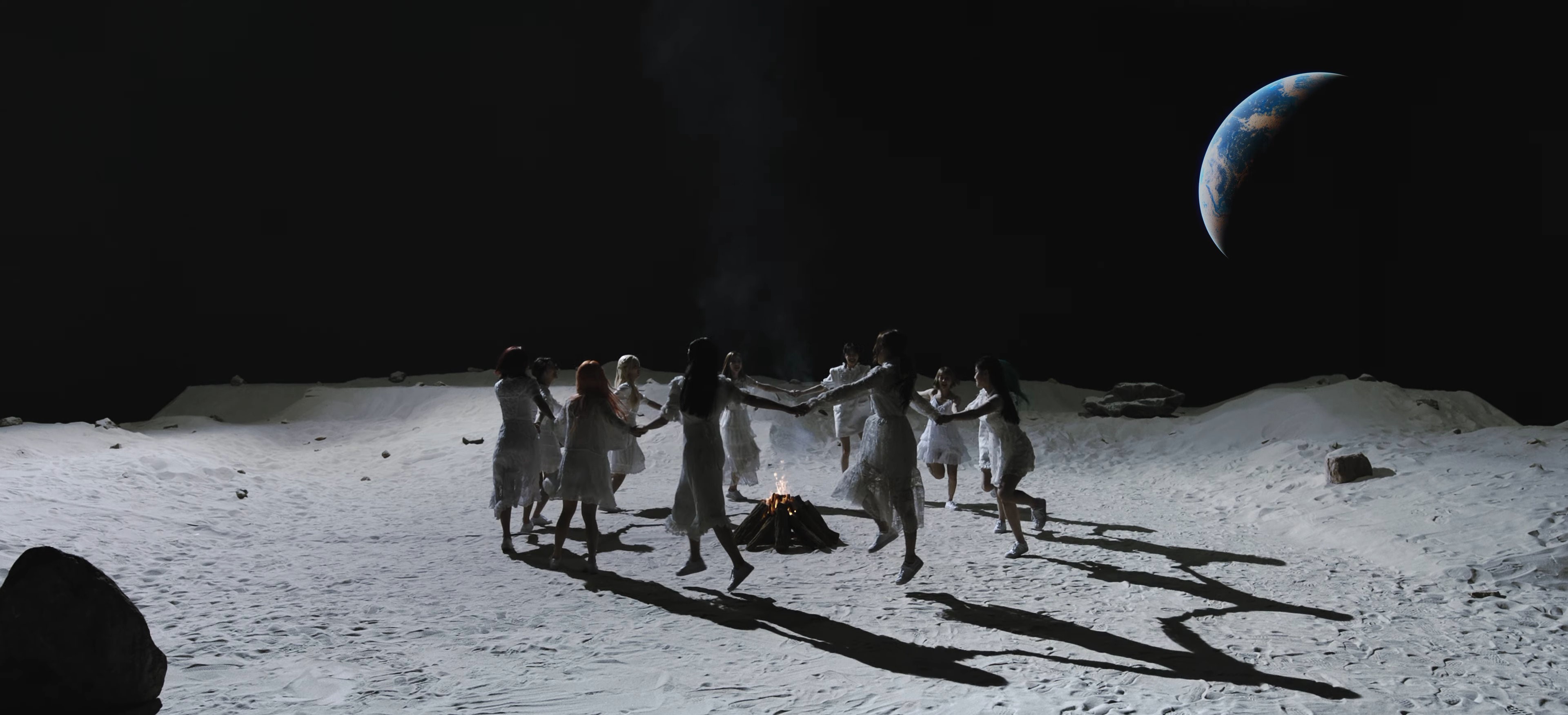 The members of LOONA dancing on the moon around a campfire, from the Why Not music video. They are wearing white dresses and are skipping in a circle, holding hands with each other. They are standing on the sandy gray surface of the moon. Black space is behind them, with the Earth floating nearby.