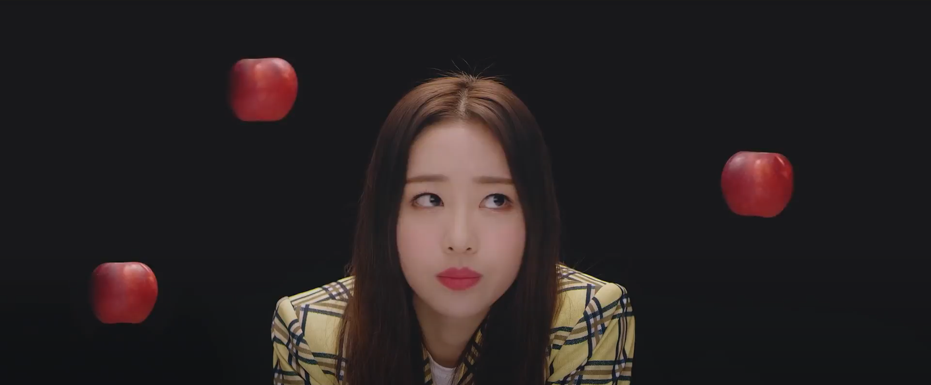 Yves looking around, surrounded by floating apples, from the New music video. She is wearing the yellow plaid yyxy uniform. She has a confused look on her face. She has long dark brown hair. There are 3 apples floating around her, all red. The background is black.