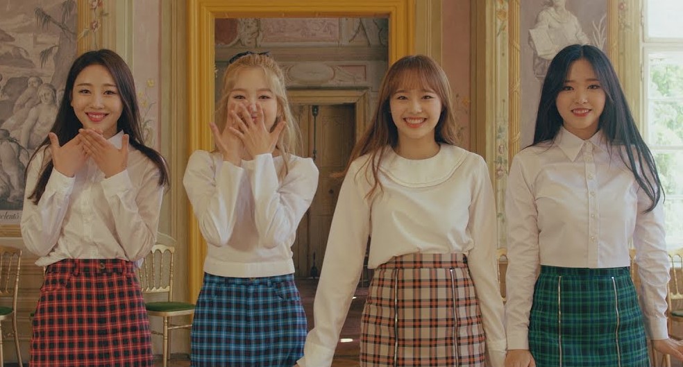 A picture of yyxy from the love4eva music video. From left to right: Yves, Go Won, Chuu, Olivia Hye. They are standing stiffly and smiling brightly, in a way that looks somewhat forced. Yves and Go Won have their hands in front of their mouths, while Chuu and Olivia's arms are down. They are wearing white shirts with plaid skirts. Yves' skirt is red, Go Won's is blue, Chuu's is peach, and Olivia's is green. Yves and Olivia have long black hair. Go Won has long blonde hair, with a black bow on the back of her head. Chuu has long light brown hair. They are standing in an extravagant golden room, with paintings and an ornate doorway behind them.