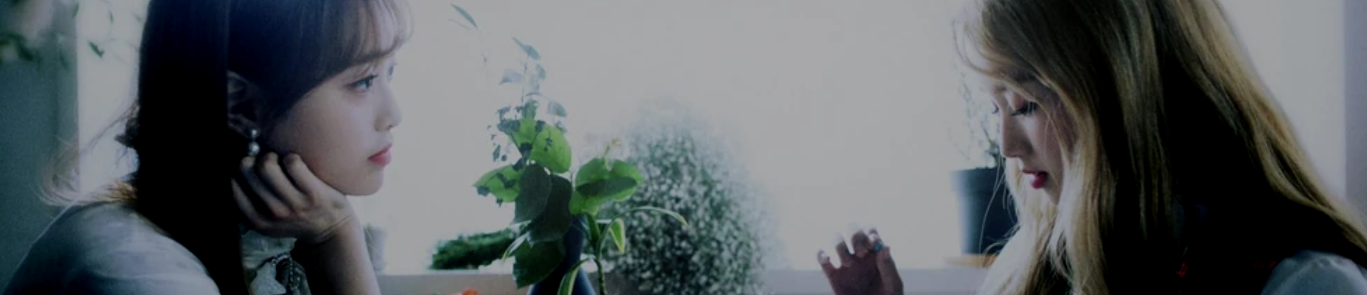 A cropped screenshot from the See Saw music video. Chuu and Go Won are sitting at a table in front of a bright window with plants in the windowsill. Chuu is on the left, looking at Go Won with her head resting in her hand. She has long brown hair and is wearing a white top. Go Won is on the right, looking down at the table. She has long blonde hair and is also wearing a white top.