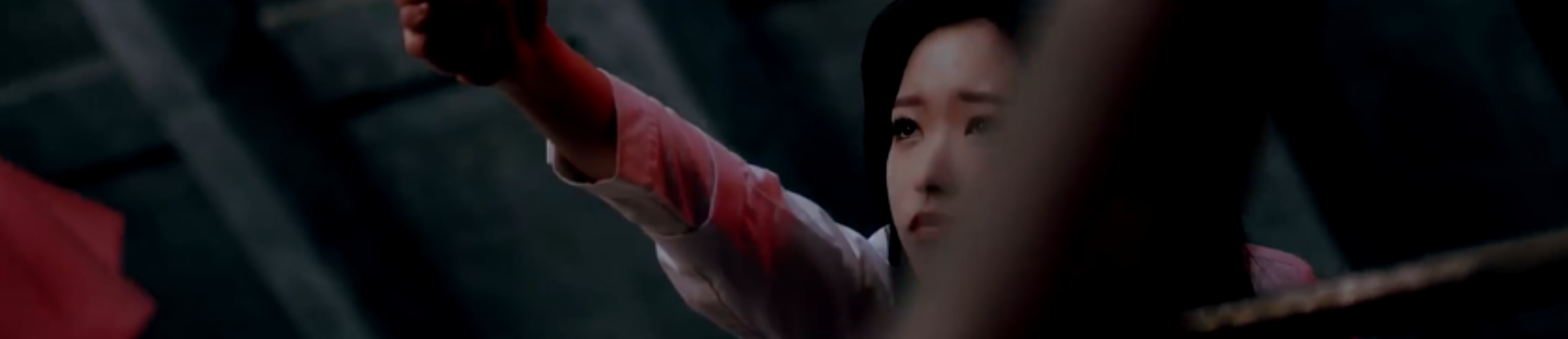 A cropped screenshot from the Eogist music video. Olivia Hye is dangling from a platform above, holding onto an unseen person's hand. She has a worried/hurt expression on her face, looking up at the other person. She is wearing a white button up shirt, and has long black hair. Behind her is an industrial looking wall. There is a railing in the foreground slightly obscuring her face.