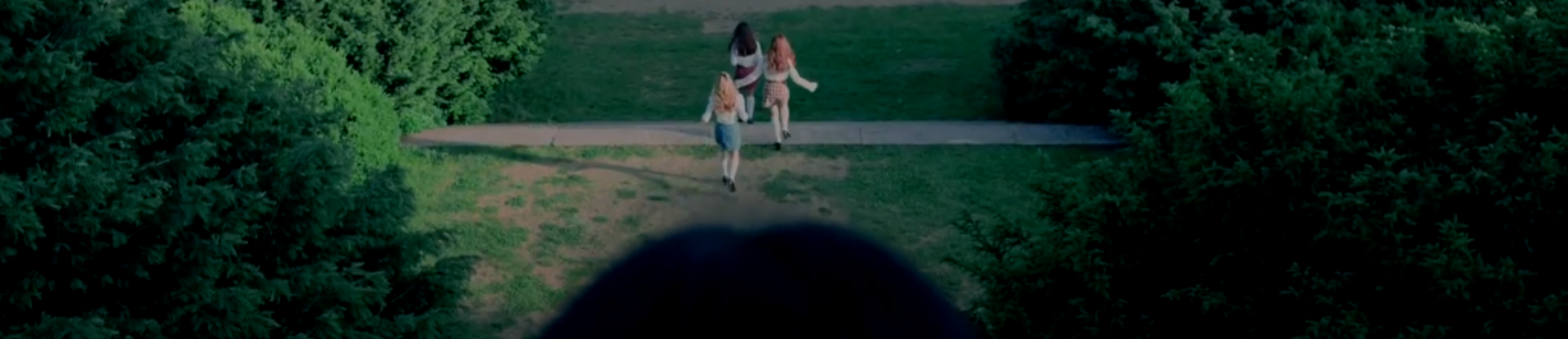 A cropped screenshot from the love4eva music video. Yves, Chuu, and Go Won are far in the background, running off in their white button up shirts and plaid skirts. They are crossing a path through green grass, surrounded by large green bushes. The back of Olivia Hye's head is in the foreground, watching them leave.