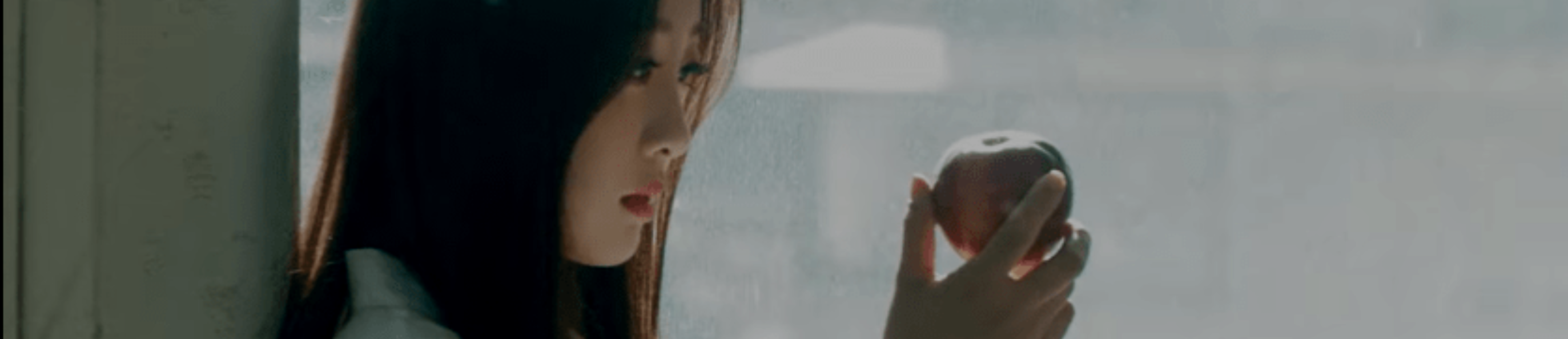 A cropped screenshot from the new music video. Yves is holding an apple in front of her, staring at it contemplatively. She is wearing a white shirt, and has long dark brown hair. She is leaned against a wall. The background is gray.