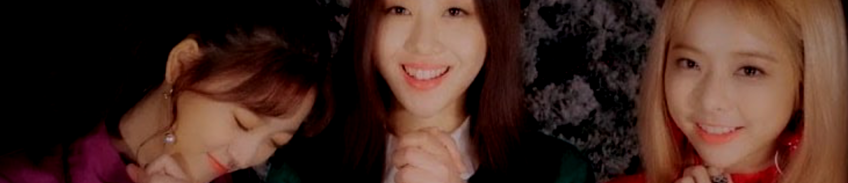 A cropped screenshot from The Carol 2.0 music video. From left to right: Choerry, Yves, Vivi. They are standing side by side in front of a gray patterned wall. Choerry has long dark brown hair and is wearing a purple top. She has her hands clasped in front of her and is tilting her head to the right, smiling with her eyes closed. Yves has long dark brown hair and is wearing a green top with a white collar. She also has her hands clasped, and is smiling towards the viewer. Vivi has long blonde hair and is wearing a red top. She is also clasping her hands and smiling at the viewer.